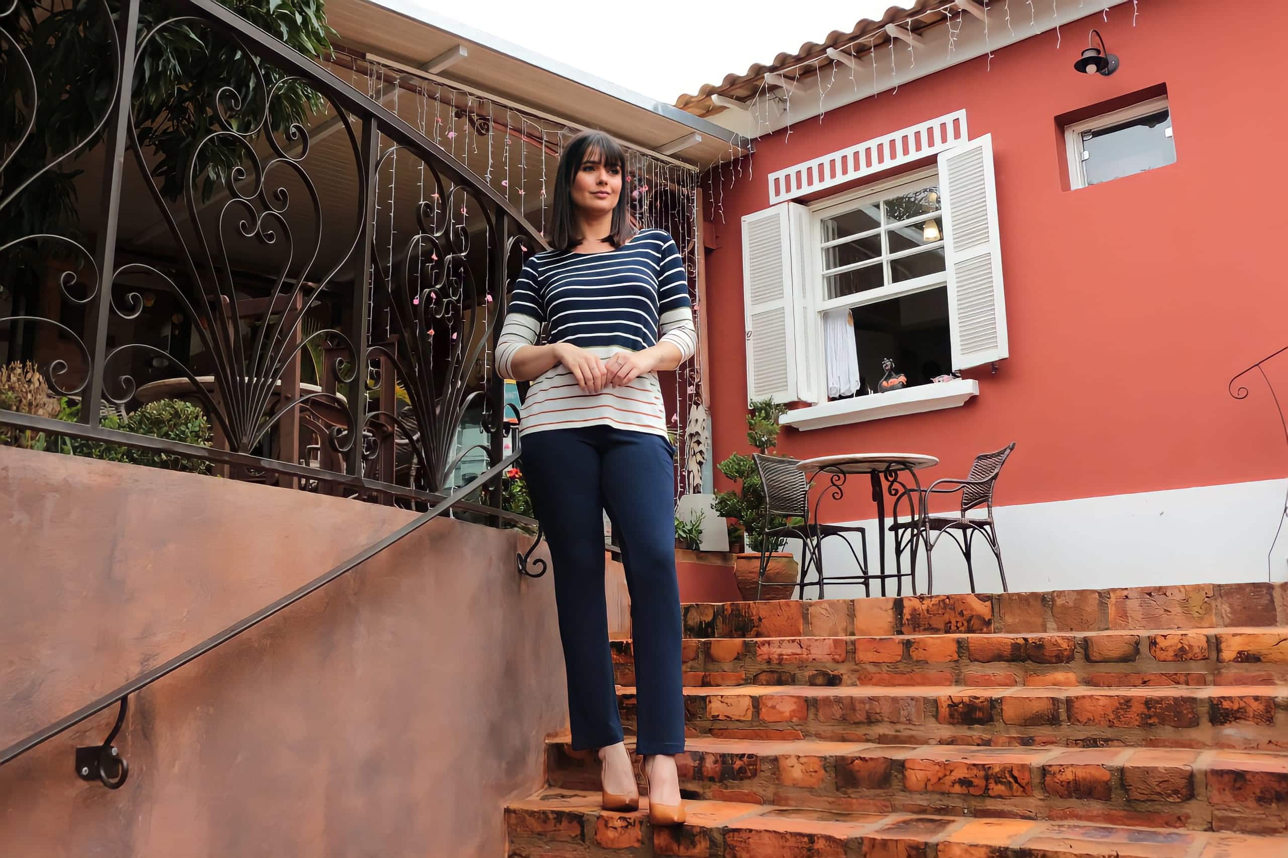 Mulher em uma casa posando para foto na escada utilizando calça escura e blusa de manga listrada
